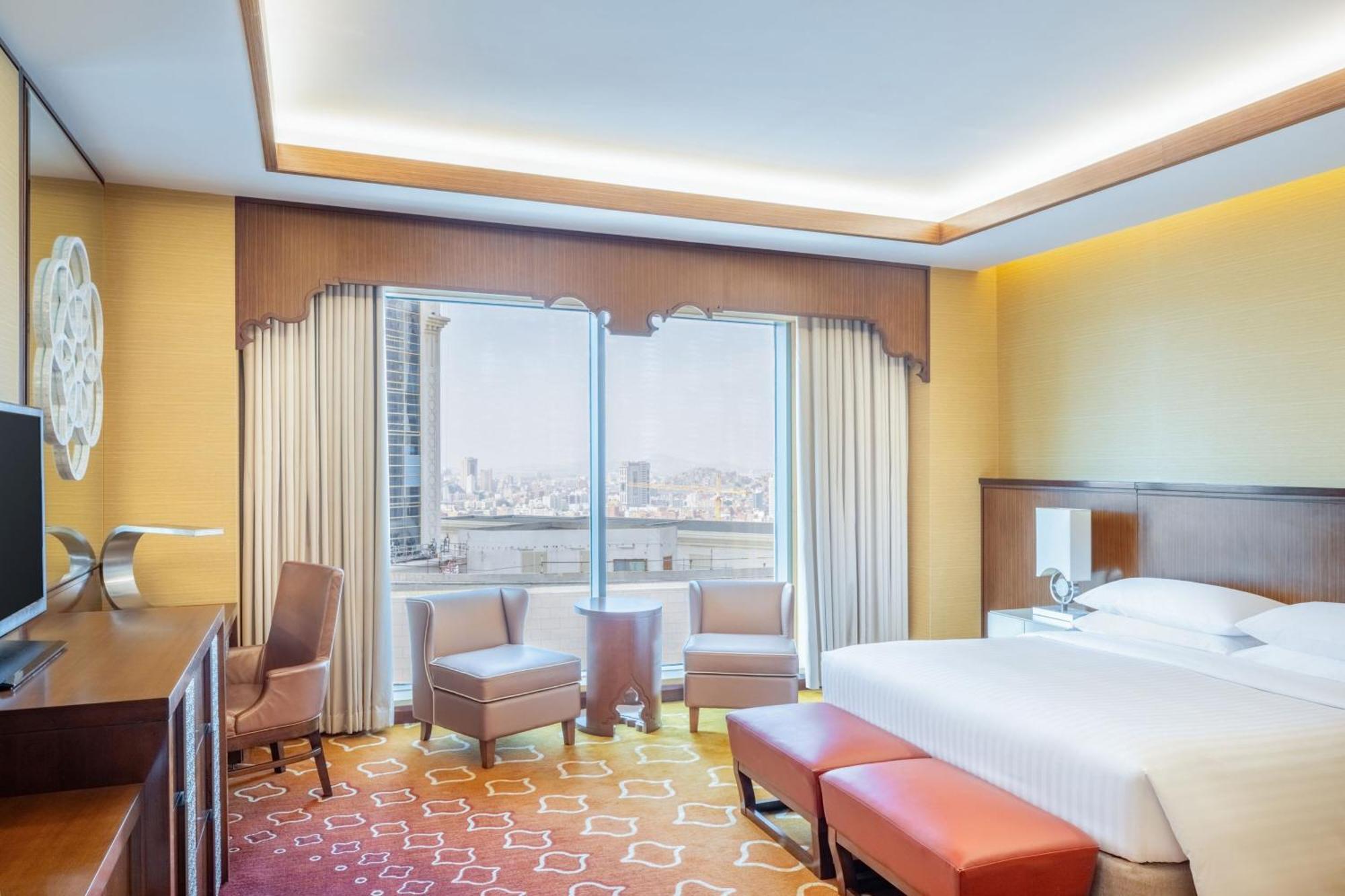 Jabal Omar Marriott Hotel Makkah Mecca Екстер'єр фото The photo shows a modern hotel room featuring a comfortable bed with crisp white linens. There are two armchairs and a small table positioned by large windows that provide a view of the city. The room has warm, inviting colors with a patterned carpet