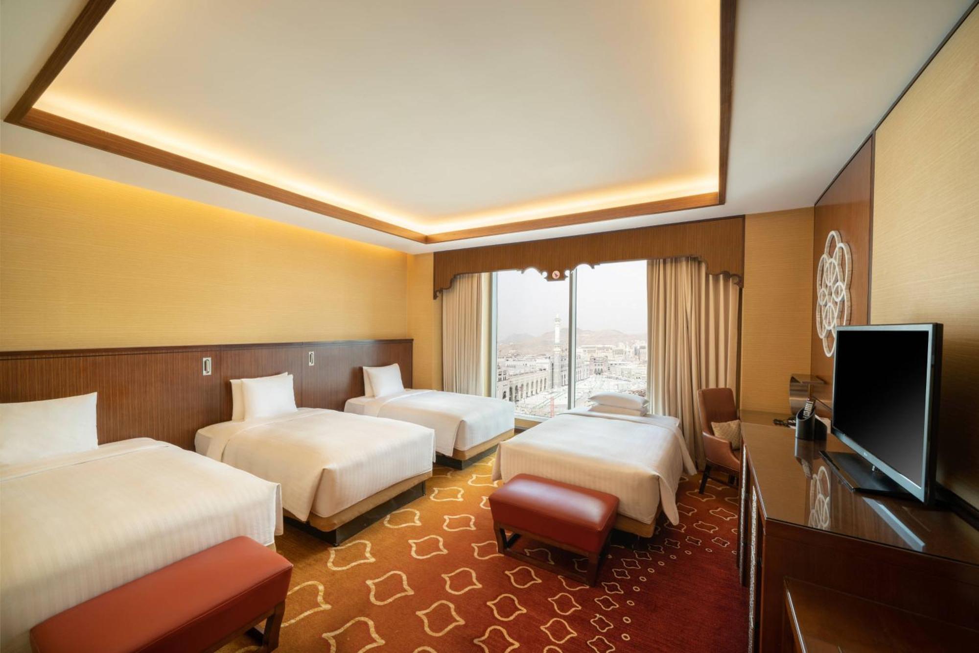 Jabal Omar Marriott Hotel Makkah Mecca Екстер'єр фото The photo shows a spacious hotel room featuring four beds arranged in a clean and modern style. The room has a warm color palette with yellow and brown tones. Natural light enters through large windows adorned with curtains, providing a view beyond. 
