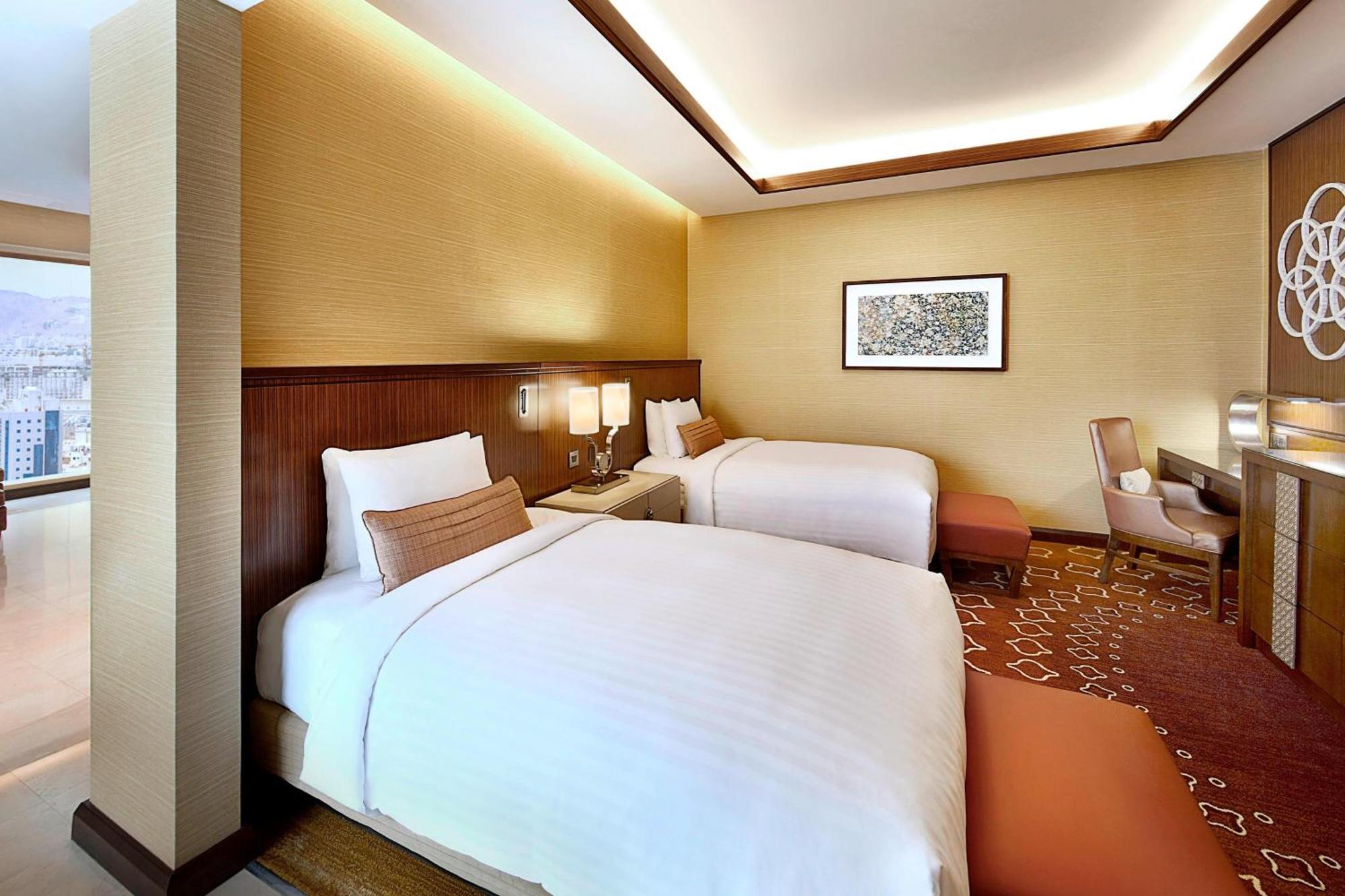 Jabal Omar Marriott Hotel Makkah Mecca Екстер'єр фото The photo shows a modern hotel room featuring two beds with white linens. The room has a warm color palette, with beige walls and wooden accents. There's a piece of artwork on the wall above the beds. A small seating area is present, along with a wor